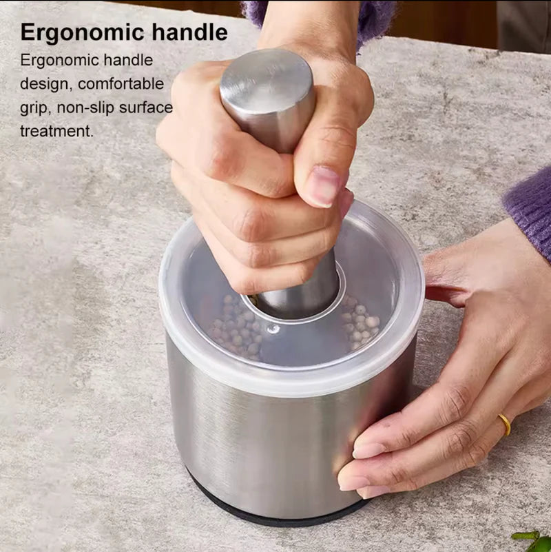 Person using a stainless steel pepper grinder with an ergonomic handle on a gray surface.