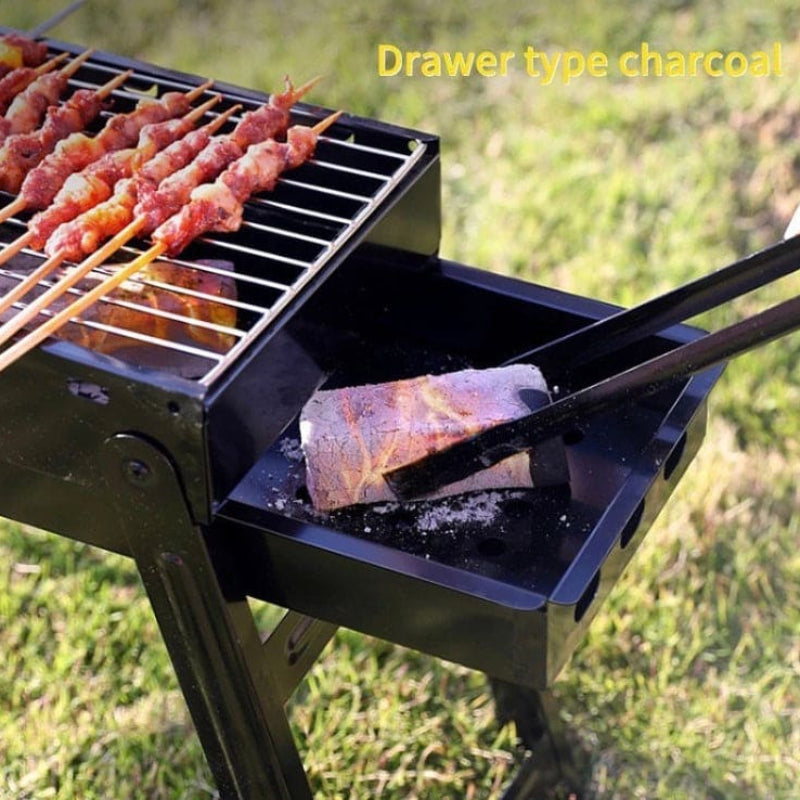 Portable charcoal grill with skewered meat and a drawer type charcoal system on a grassy background.